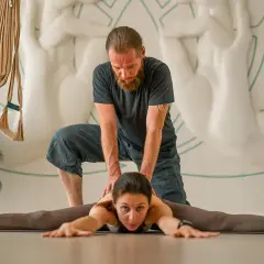 Instruktor pomaga kobiecie pogłębić pozycję Upavistha Konasana, rozciągając nogi w szerokim rozkroku.