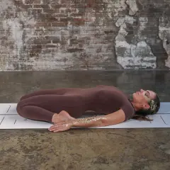 Kobieta w pozycji supta virasana na macie. Jej ciało jest wygięte do tyłu, nogi zgięte w kolanach, stopy pod pośladkami.