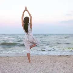 Kobieta w pozycji drzewa na plaży, z rękami złożonymi nad głową. Spokojne morze i pastelowe niebo tworzą tło.