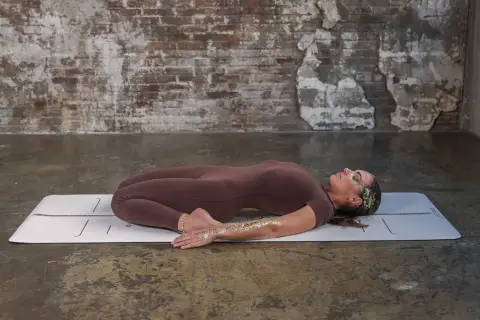 Kobieta w pozycji supta virasana na macie. Jej ciało jest wygięte do tyłu, nogi zgięte w kolanach, stopy pod pośladkami.