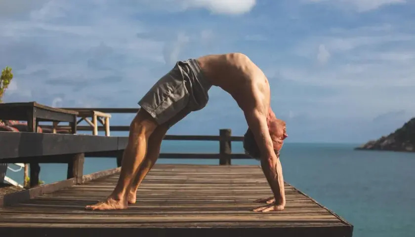 Mężczyzna wykonuje pozycję urdhva dhanurasana na drewnianym tarasie z widokiem na ocean i niebo.