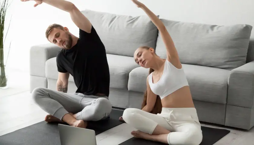 Para ćwiczy jogę w pozycji sukhasana, wykonując skłony boczne, patrząc na laptopa.