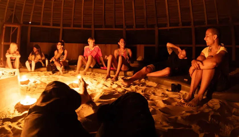 Grupa ludzi siedzi na piasku w okrągłej, drewnianej chacie, otoczona świecami. Rozmawiają i śmieją się, przygotowując się do ceremonii. Ayahuasca cena może być różna, ale to doświadczenie jest bezcenne.