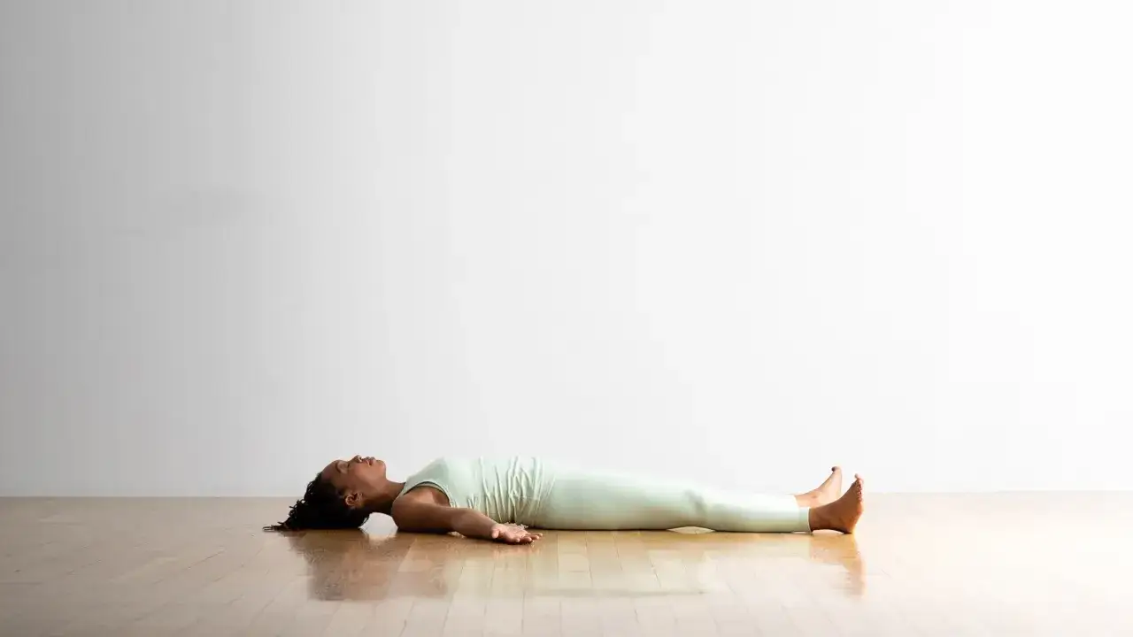 Kobieta w jasnym stroju do jogi leży na podłodze w pozycji savasana, odpoczywając po sesji.